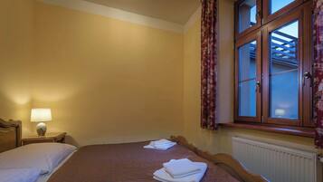 A hotel room with two beds, a nightstand with a lamp, a window with curtains, and a radiator.