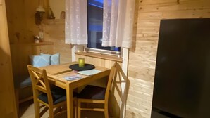 Ein kleiner Holzraum mit Tisch, zwei Stühlen, einem Fenster mit weißen Vorhängen, einem schwarzen Kühlschrank und einer Holzwand.