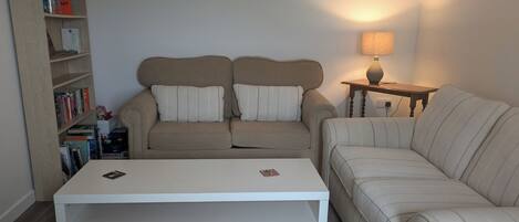 Uma sala de estar com sofá, mesa de centro, mesa lateral com abajur e estante de livros.
