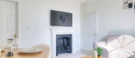 A living room with a fireplace, a television mounted above it, a sofa, and a dining table set with plates and glasses.