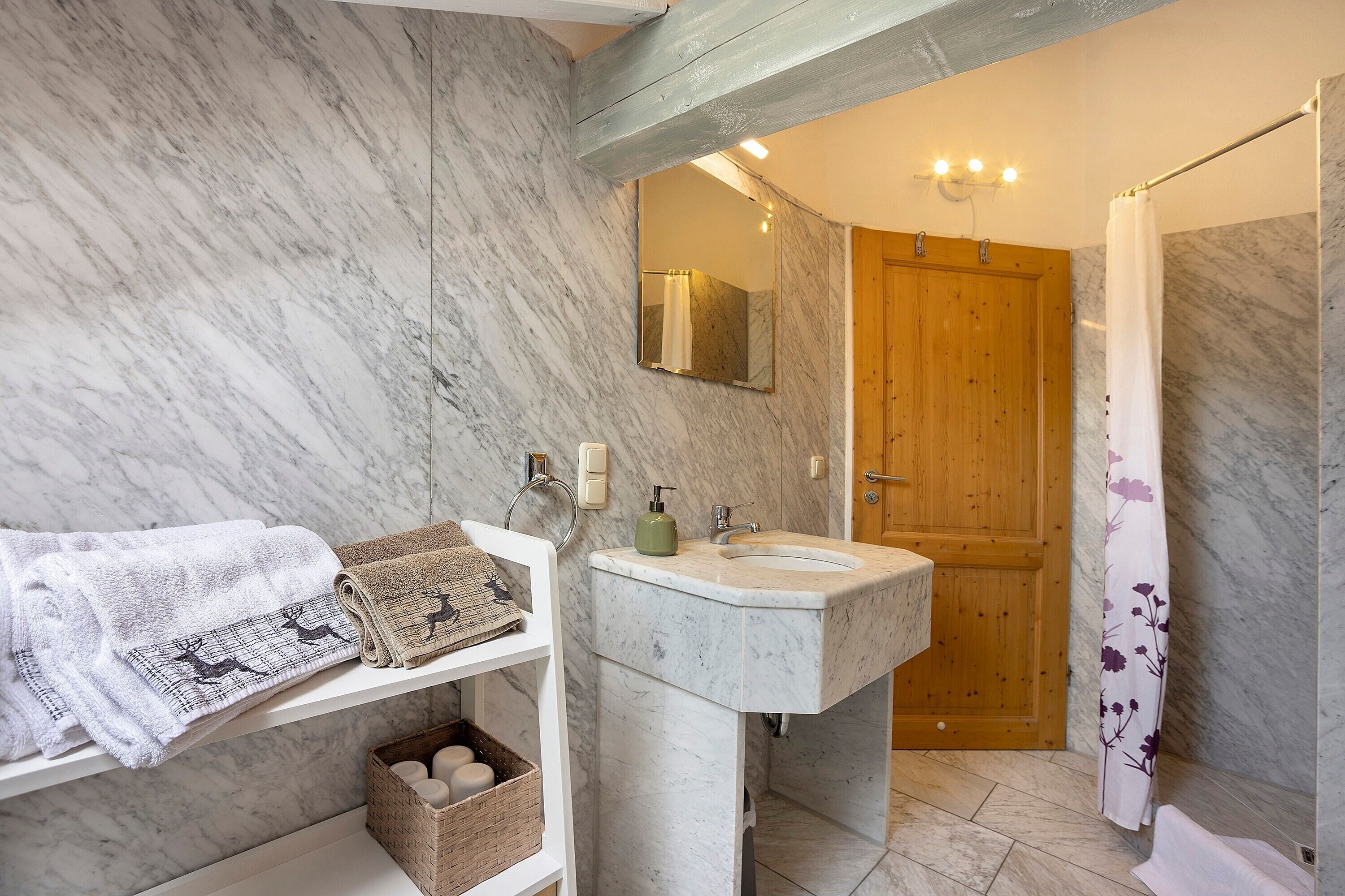 A bathroom with a marble wall, wooden door, sink, and a shelf with towels.