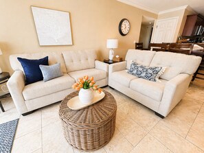 A living room with two sofas, a wicker coffee table, a wall clock, and a kitchen in the background. - Front Row to the Main Channel - 3BR walk in unit! (Osage Beach)