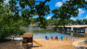 A sandy beach with a playground, including a slide and swings, and a row of colorful Adirondack chairs facing a lake with boats and houses in the background. - Front Row to the Main Channel - 3BR walk in unit! (Osage Beach)