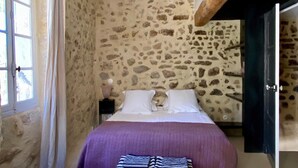 A bedroom with a stone wall, a bed with white pillows, a purple bedspread, a bench, a white chair, and a window with curtains. - Holiday Home 'Présbytère' with Shared Pool, Shared Garden and Wi-Fi (Lieuran-lès-Béziers)