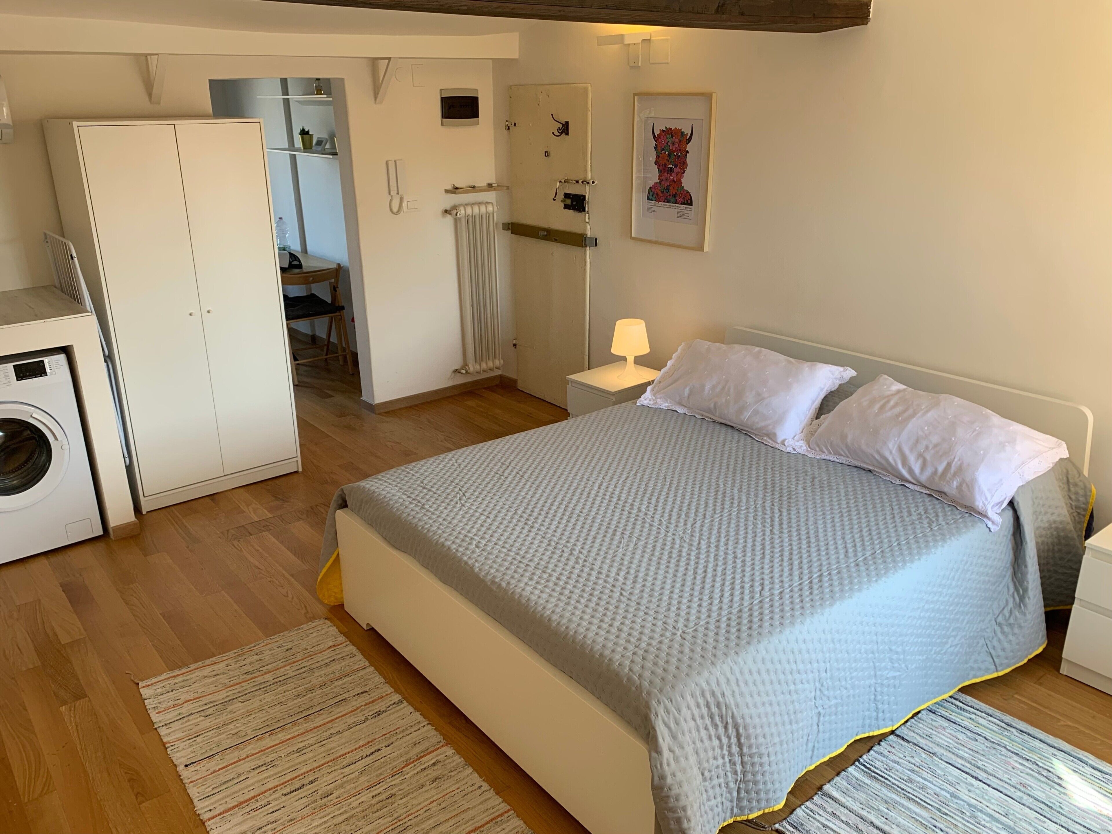 A bedroom with a bed, a washing machine, a radiator, a nightstand, and a framed picture on the wall.