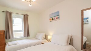 Une chambre avec deux lits, une fenêtre avec des rideaux, une lampe de chevet, un tableau représentant une scène de plage et un miroir sur le mur.