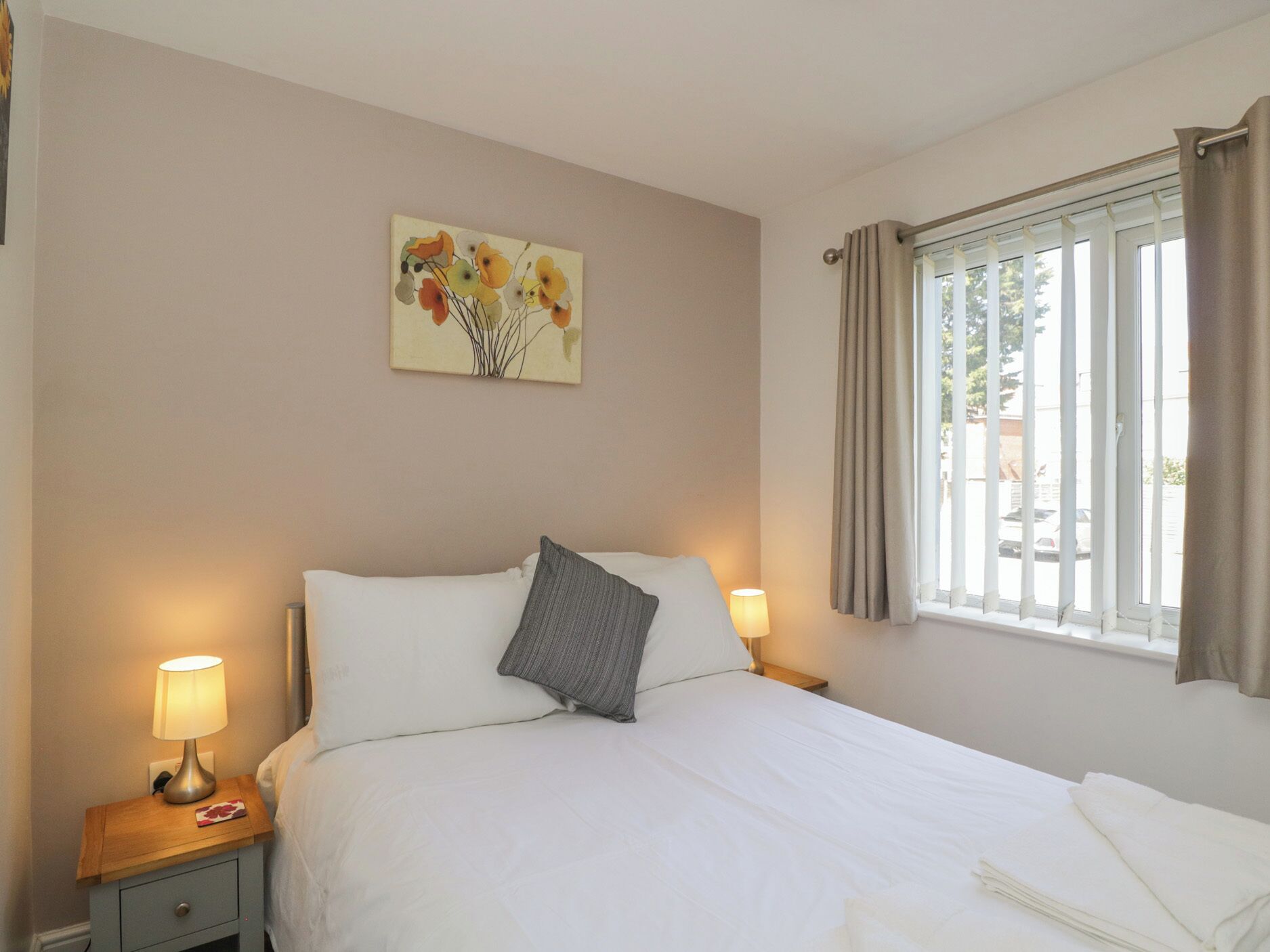 A hotel room with a bed, bedside lamps, a window with curtains, and a floral painting on the wall.