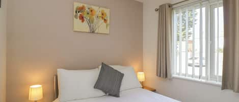 A hotel room with a bed, bedside lamps, a window with curtains, and a floral painting on the wall.