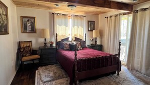 A bedroom with a wooden ceiling, a four-poster bed, two bedside tables with lamps, a chair, and a large window with curtains.