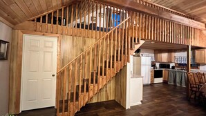 A wooden staircase leading to an upper level, a kitchen area with a refrigerator and microwave, and a dining area with chairs.
