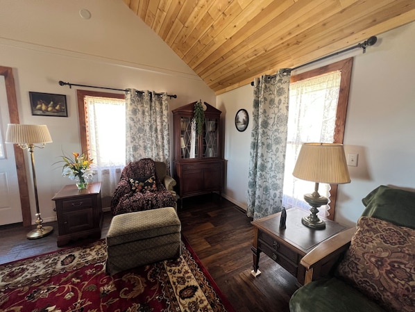 Una sala de estar acogedora con techo de madera, un sillón estampado, un pequeño reposapiés, un armario y una alfombra floral.