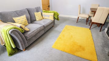 Un soggiorno con un divano grigio, cuscini gialli e verdi, un tappeto giallo e una porta in legno sullo sfondo.
