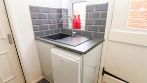 A compact kitchen with a built-in sink, a small refrigerator, and a window with a view of a brick wall. - 17 Fairfield Road (Newcastle-upon-Tyne)