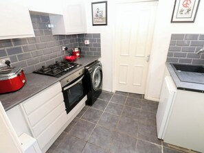 A compact kitchen with a gas stove, a red kettle, a washing machine, and a white door. - 17 Fairfield Road (Newcastle-upon-Tyne)