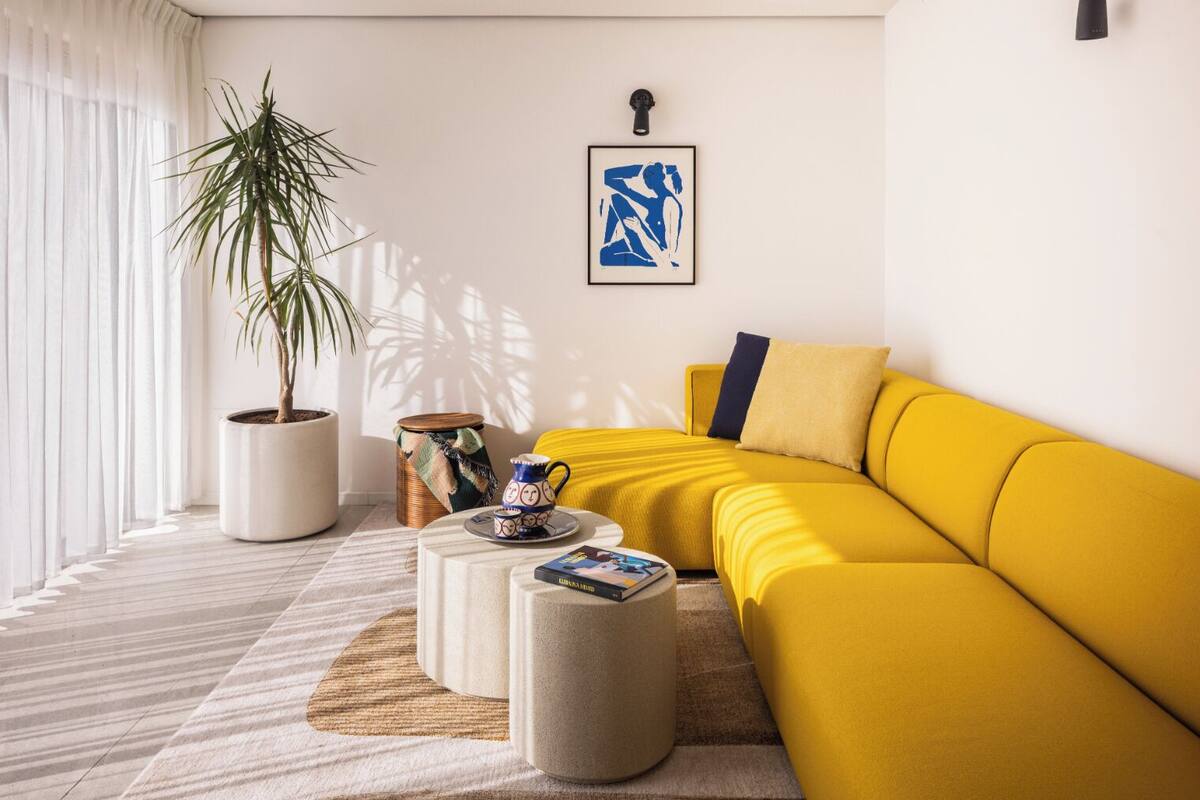 Ein modernes Wohnzimmer mit einem gelben Sofa, einer Topfpflanze, einem Couchtisch und einem gerahmten Kunstwerk an der Wand.