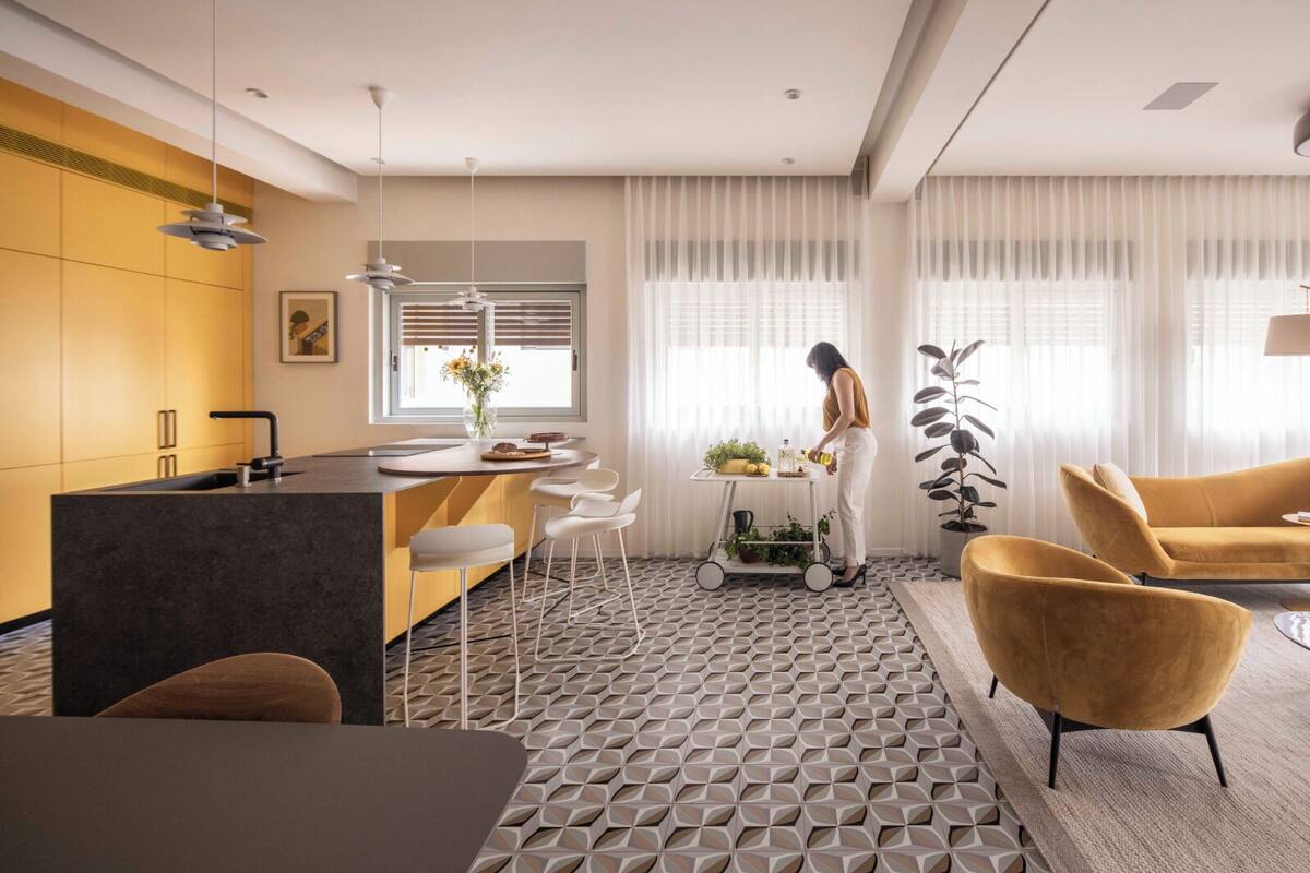 A modern kitchen with a central island, white bar stools, a dining table, and a living area with yellow chairs.