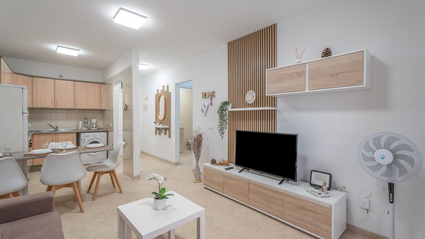 A modern living room with a TV, white furniture, a dining area, and a kitchen in the background. - Vacation Apartment 'Vivienda Carolina' with Mountain View, Shared Pool and Wi-Fi (Santa Cruz de Tenerife)