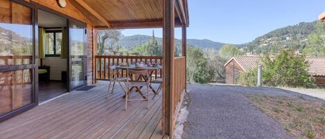 A wooden deck with a table and chairs, a sliding glass door leading to a room, and a view of a hilly landscape.