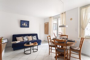 Ein Wohnzimmer mit blauem Sofa, einem Esstisch mit Stühlen, einem gerahmten Bild an der Wand und einem Fenster mit Vorhängen.