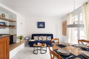 Un salon moderne avec un canapé bleu, une table à manger dressée pour quatre personnes et un coin cuisine doté d’étagères en bois.