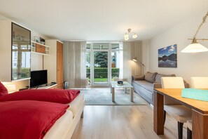 A modern living room with a sofa, a TV, a dining table, and a large window offering a view of greenery. - Elbappartement Dresden – Modern Apartment Near the Elbe with Terrace and Garden Access (Dresden)