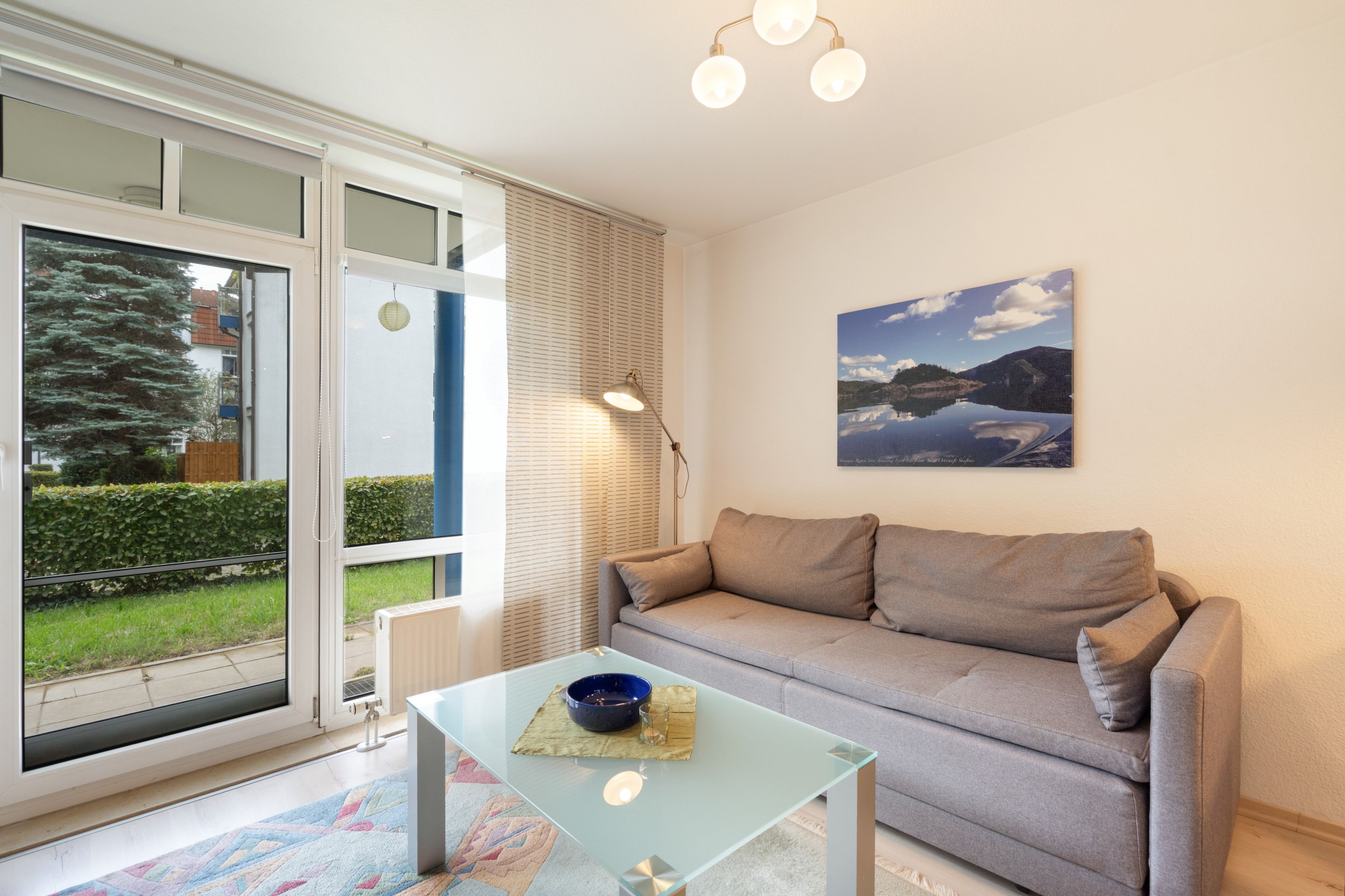 A modern living room with a glass coffee table, a grey sofa, a painting of a lake, and a view through a sliding glass door.