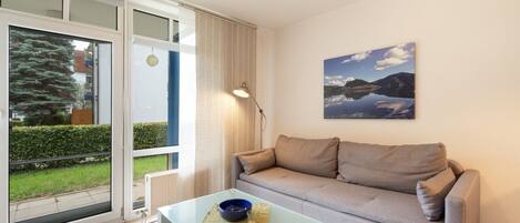 A modern living room with a glass coffee table, a grey sofa, a painting of a lake, and a view through a sliding glass door.