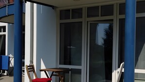 A covered outdoor seating area with a table and two chairs, a window, and a blue pillar. - Elbappartement Dresden – Modern Apartment Near the Elbe with Terrace and Garden Access (Dresden)