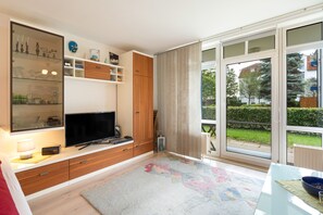 A modern living room with a flat-screen TV, wooden cabinets, and a large window with blinds. - Elbappartement Dresden – Modern Apartment Near the Elbe with Terrace and Garden Access (Dresden)