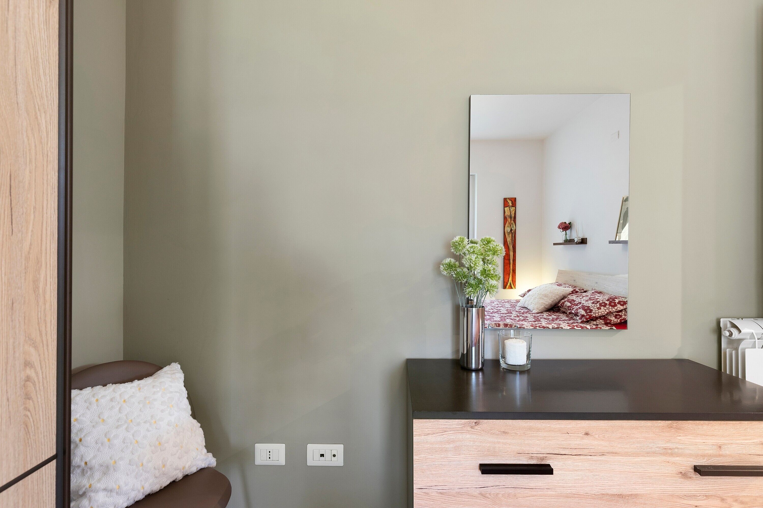 Une chambre à coucher moderne avec une commode en bois, une table de chevet ornée d’un vase et d’une bougie, et un grand miroir qui reflète la pièce.