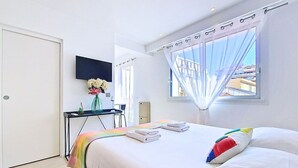 A hotel room with a bed, a desk, a chair, a TV, and a window with a view of buildings. - N&J - STUDIO ANNA - Central Nice • AC • Walk to beach (Nice)