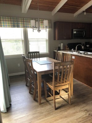 Een eetgedeelte met een houten tafel en stoelen, een keuken met donkere kastjes, en een raam met jaloezieën.
