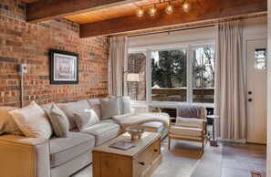 Ein gemütliches Wohnzimmer mit Backsteinwand, einer Couch, einem Couchtisch, einem Sessel und einem Fenster mit Vorhängen.