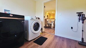A laundry area with a front-loading washer and dryer, a vacuum cleaner, and a dining area visible through an open door. - LUX Presbyterian Mercy Acrisure
PNC
PPG
31st Street Studios (Pittsburgh)