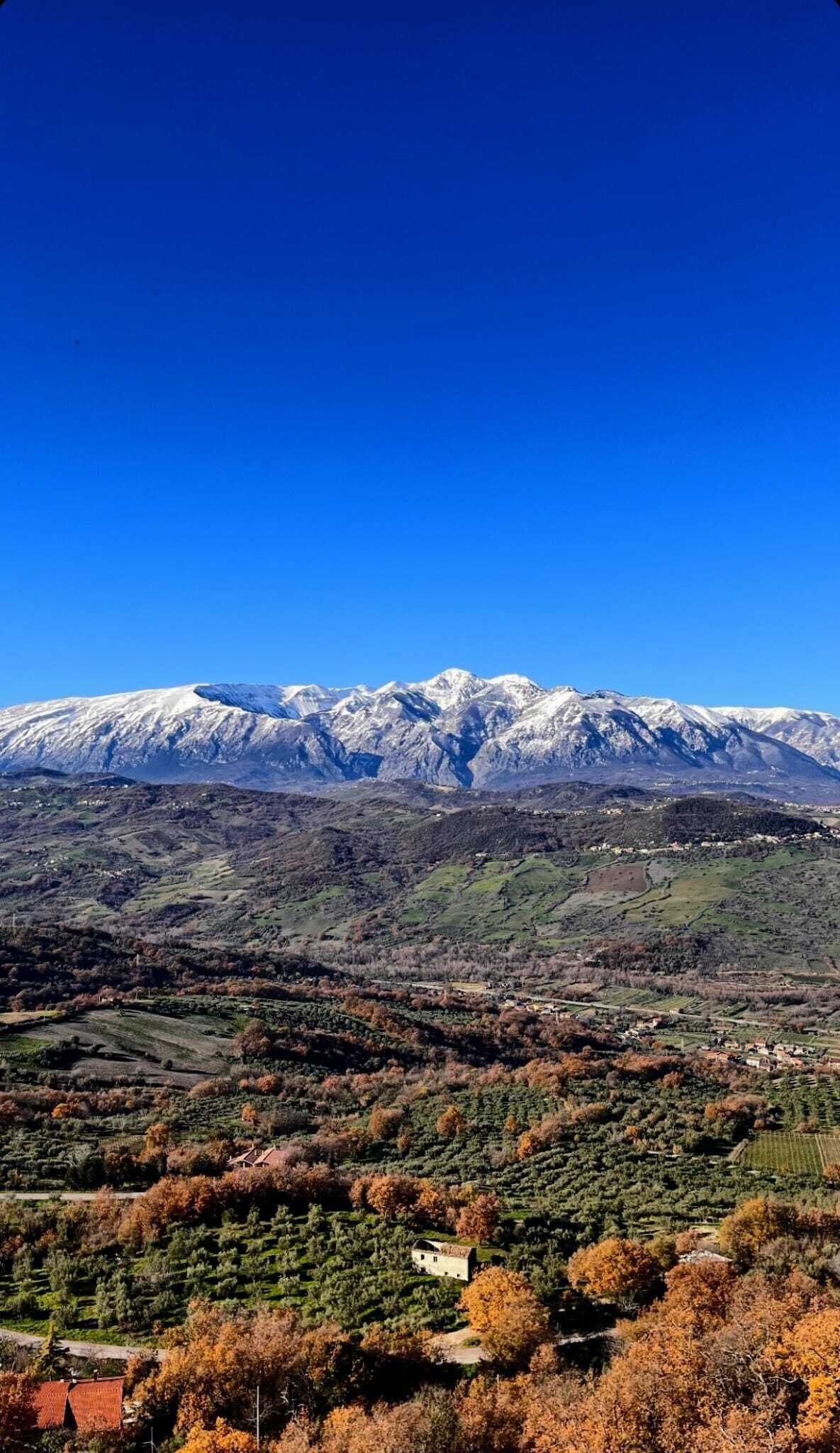 Paesaggio montuoso con cime innevate, una valle punteggiata da edifici e un cielo azzurro intenso.