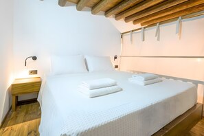 Una cama bien hecha con sábanas y toallas blancas, una mesita de noche de madera con una lámpara y un travesaño de madera en el techo.