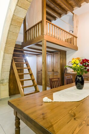 Una escalera de madera lleva a un dormitorio en el altillo, con barandilla de madera. En primer plano, se ve una mesa de comedor con sillas y un jarrón de flores.