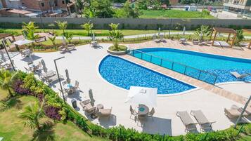 Piscina com espreguiçadeiras e mesas, rodeada por vegetação e vista para a cidade.