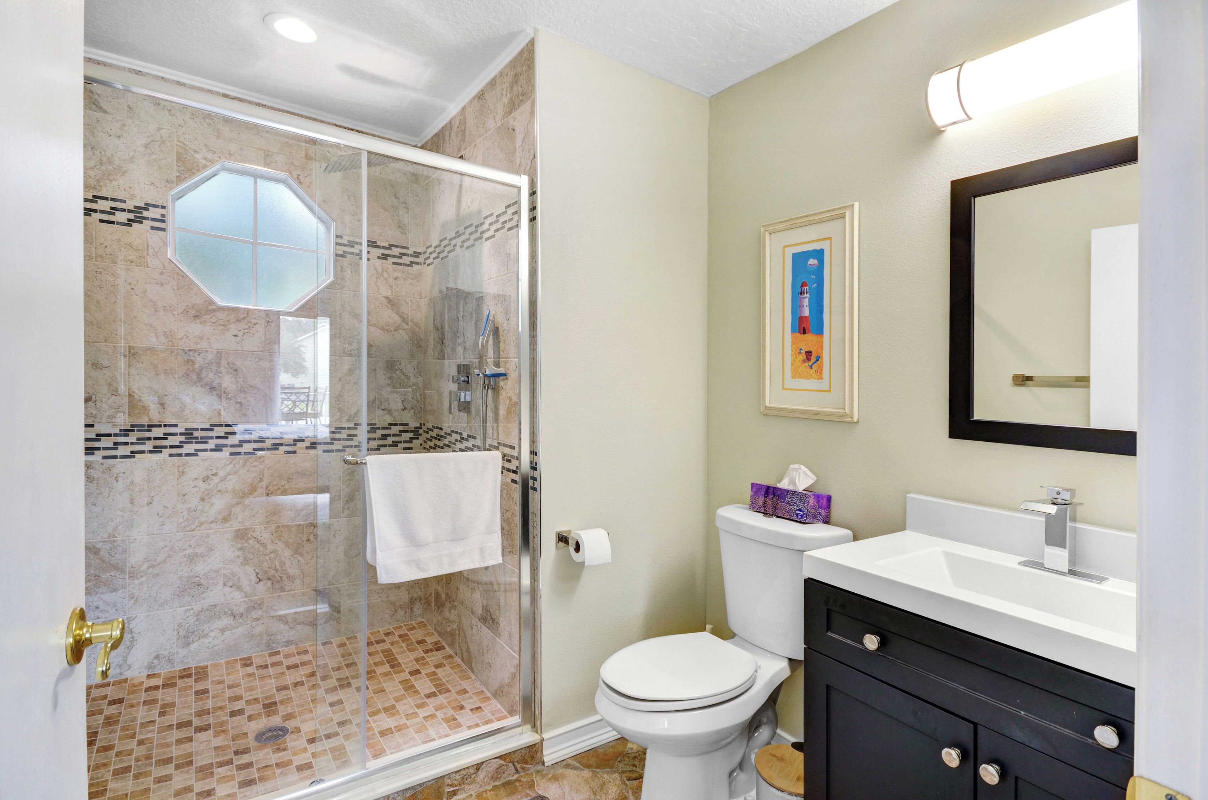 A bathroom with a glass-enclosed shower, a toilet, a sink with a mirror, and a framed picture on the wall.
