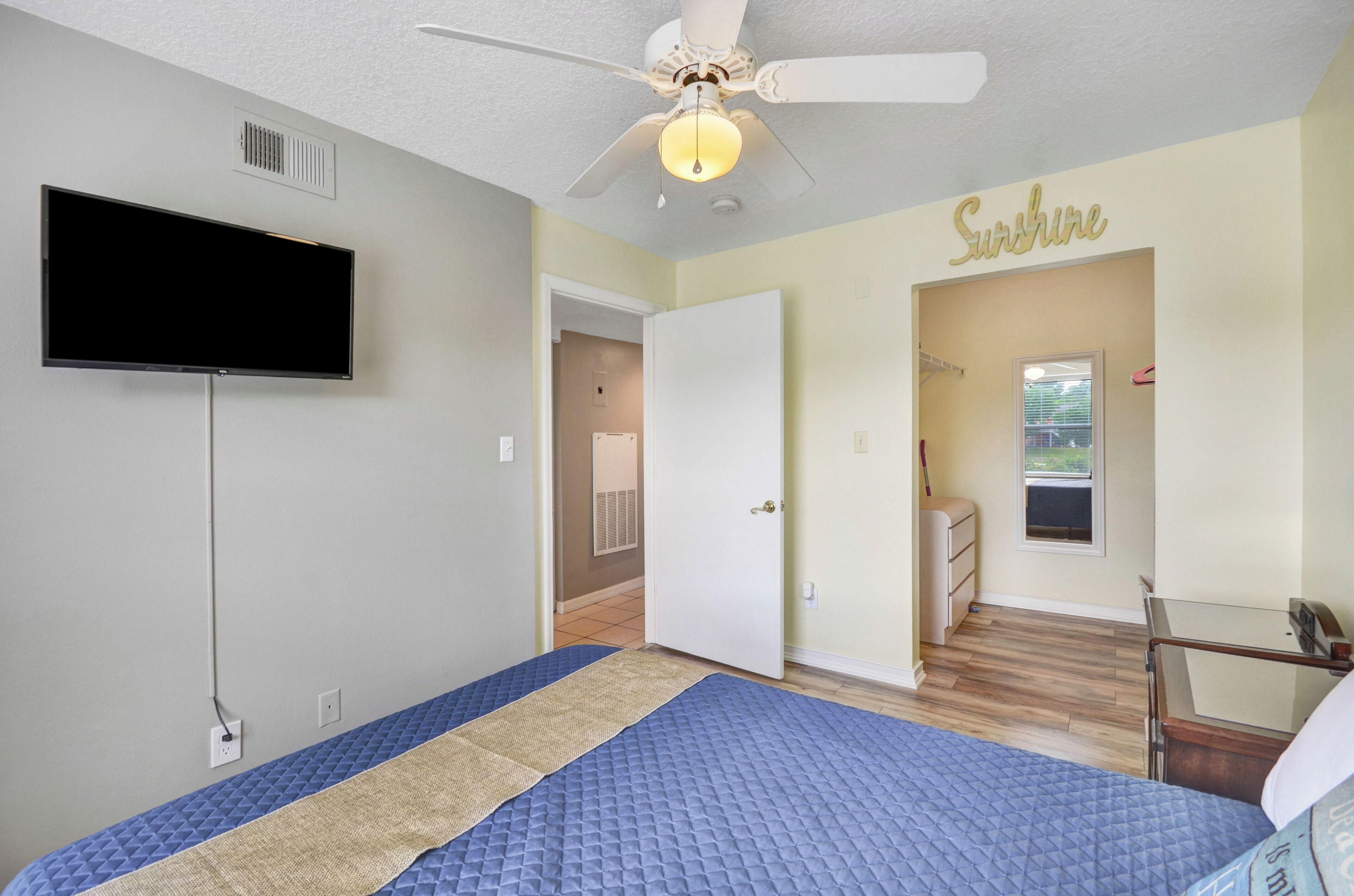 A bedroom with a bed, a ceiling fan, a TV mounted on the wall, and a door leading to another room.