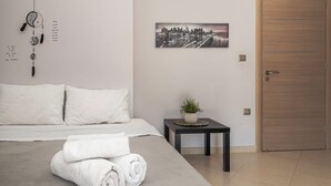 Ein ordentlich bezogenes Bett, gerollte weiße Handtücher, ein Stadtbild an der Wand, ein kleiner Tisch mit einer Pflanze und eine Holztür.