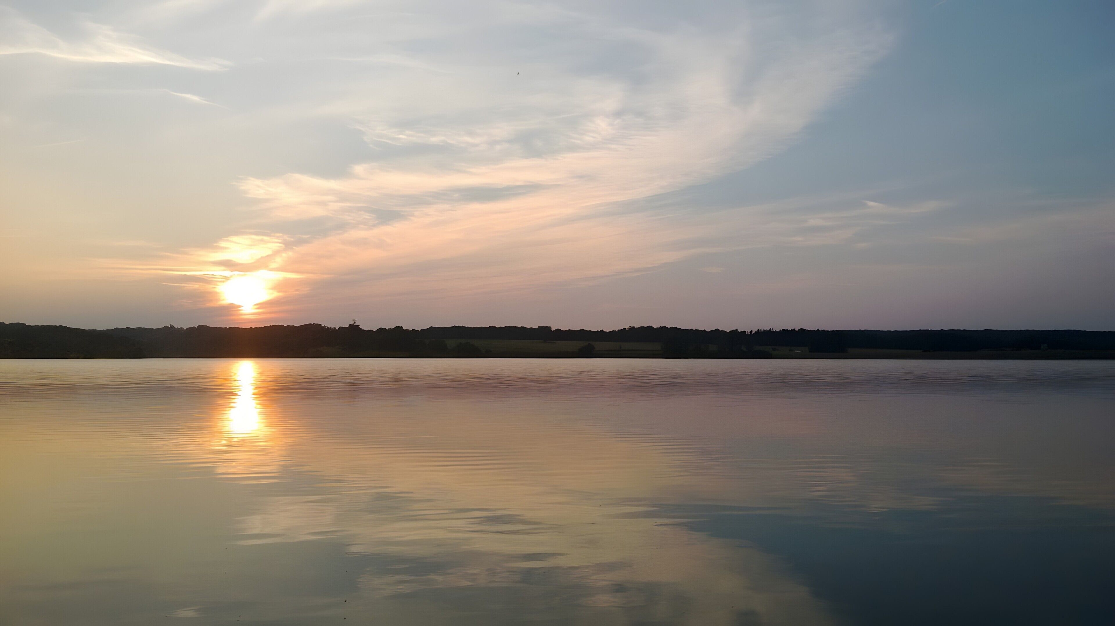 A calm lake with a sunset reflecting on the water, a distant tree line, and a clear sky.