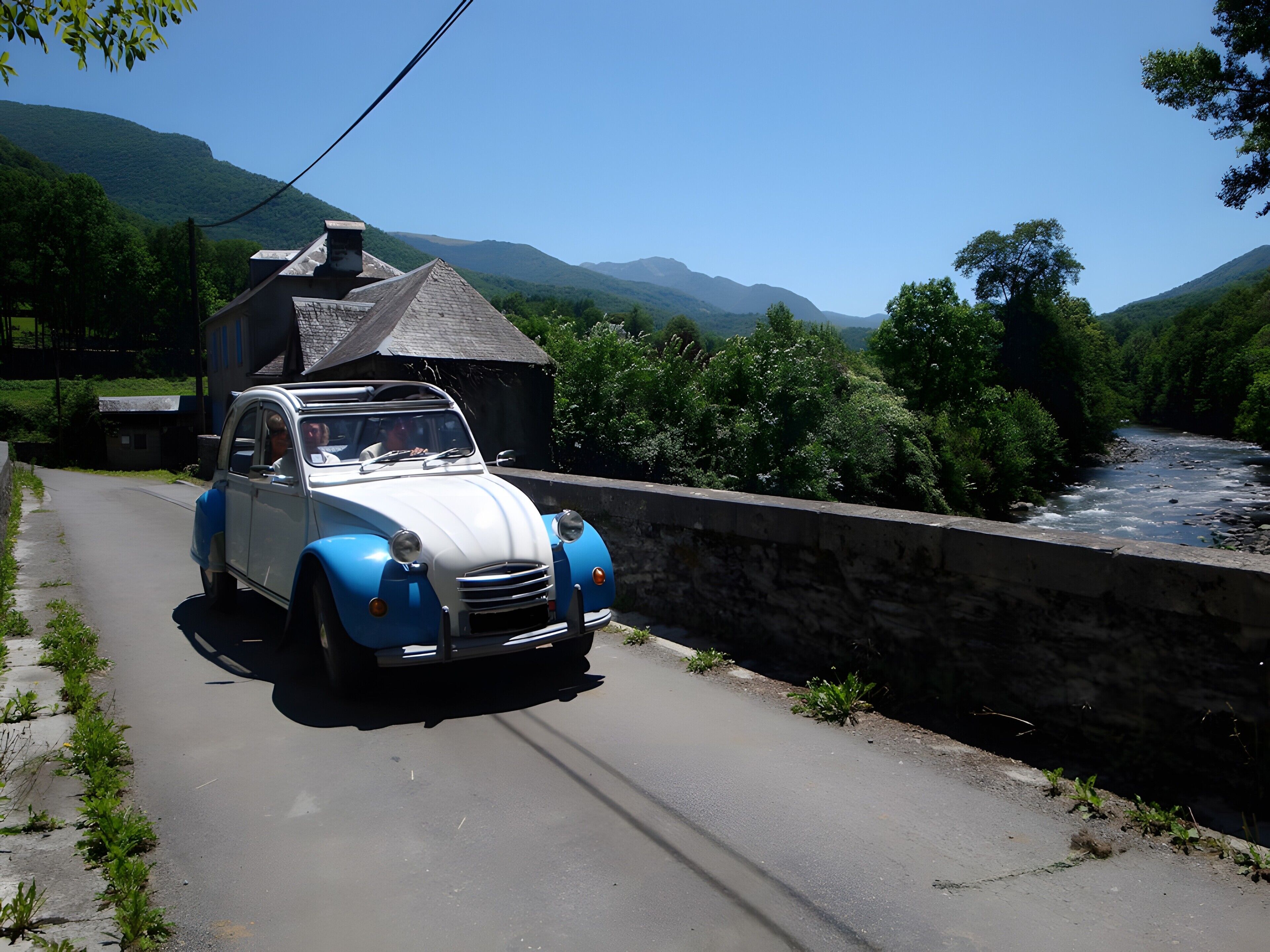 Un coche antiguo circulando por un camino estrecho junto a un río, con montañas al fondo.