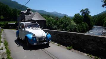 Un coche antiguo circulando por un camino estrecho junto a un río, con montañas al fondo.
