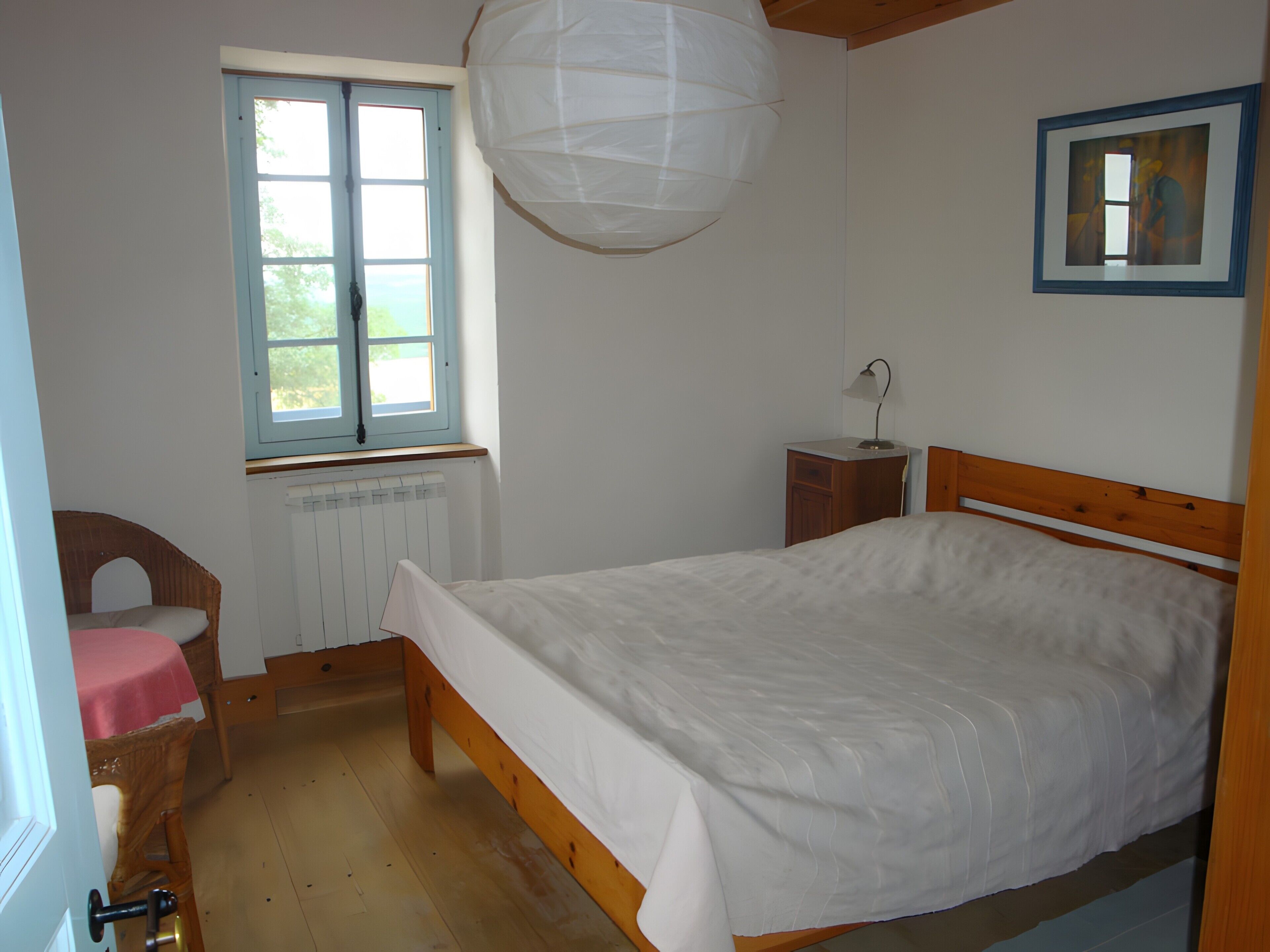 Ein Schlafzimmer mit einem hölzernen Bett, einem Fenster, einem Heizkörper, einem Sessel und einem Nachttisch.
