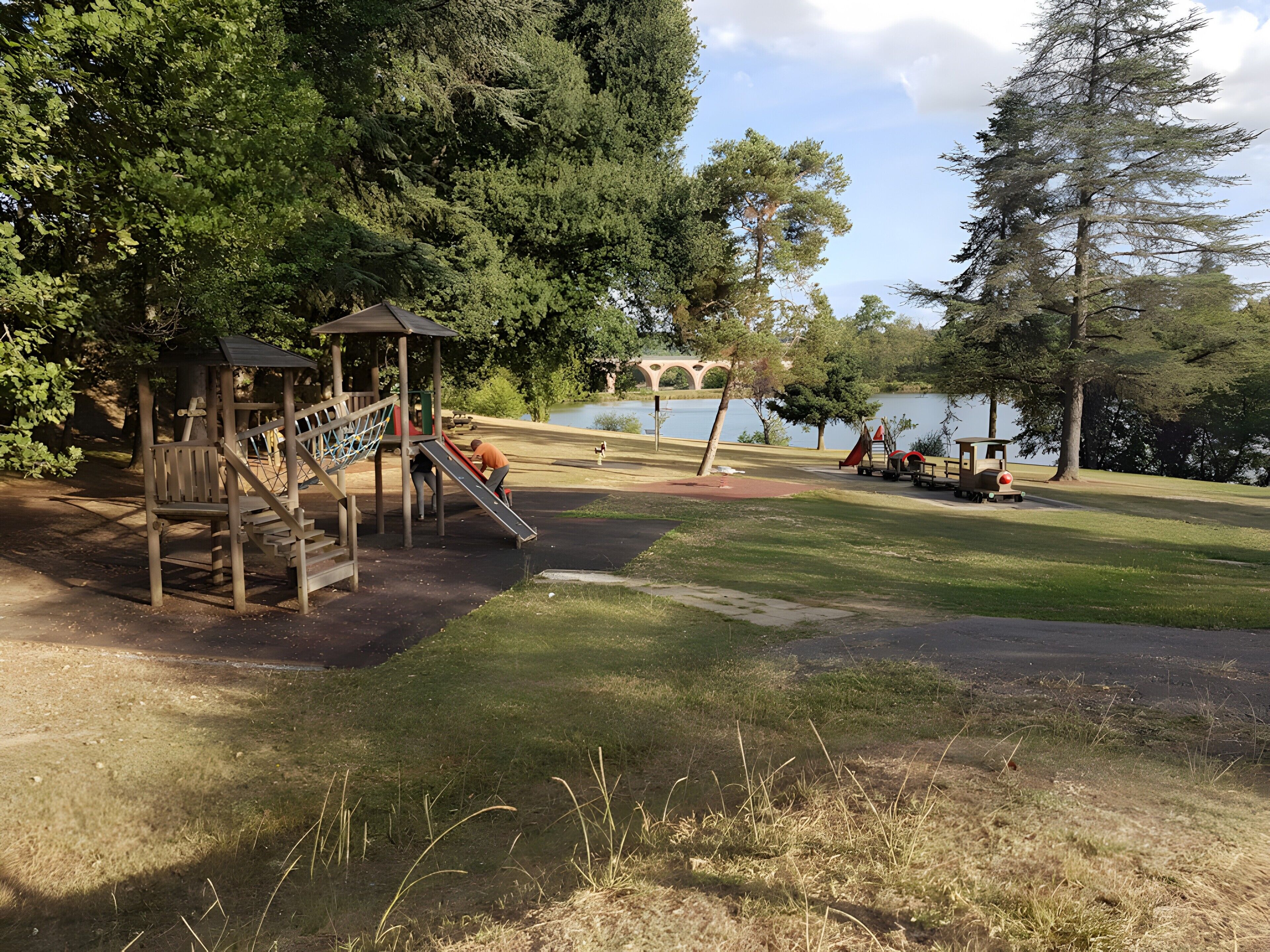 A playground with a slide and climbing structure, a small train, and a lake in the background.