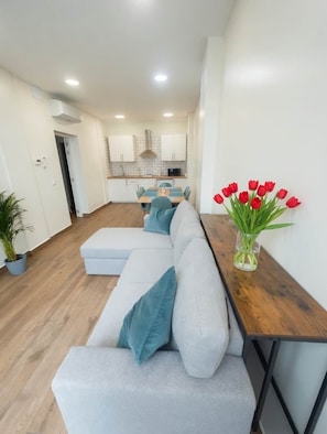 Un salon moderne avec un canapé gris, une table d’appoint en bois sur laquelle est posé un vase de tulipes rouges, et une cuisine en arrière-plan.