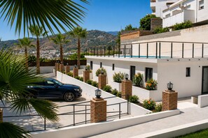 Un bâtiment moderne à deux étages, avec une voiture garée devant, entouré de palmiers et offrant une vue sur un paysage vallonné.