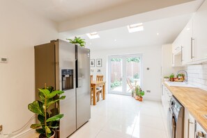 A modern kitchen with a built-in refrigerator, a wooden dining table, and a sliding glass door leading to a garden. - Charming Garden Home with Free Parking (Morden)