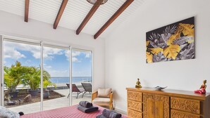 Un dormitorio con una cama grande, un armario de madera, una silla y vista al océano a través de una puerta corrediza.
